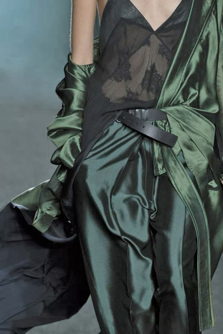 a woman is modeling a green dress with a black lace top on a fashion runway. the dress has a flowing silhouette and a black belt around the waist. the model's hair is pulled back and she is wearing minimal makeup.