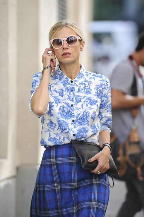 a blonde woman in a blue and white patterned shirt and a blue plaid skirt is seen outdoors. she's wearing pastel pink sunglasses and a black clutch. her style is casual yet elegant.  she appears to be in a city setting.  there are blurred figures of other people in the background.