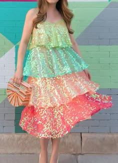 a young woman in a colorful sequin dress is posing against a brick wall. she is holding a colorful clutch bag. the dress is tiered and the colors are vibrant. she is smiling and her hair is flowing.  she looks like she is having fun and is ready for a night out.