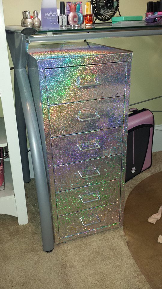 the image shows a six drawer metal cabinet with silver handles and a glittery finish. it is sitting next to a desk with various cosmetics and nail supplies on top, including nail polish remover, nail polish bottles, and makeup. a pink bag is next to the cabinet, and the image is taken on a carpeted floor.