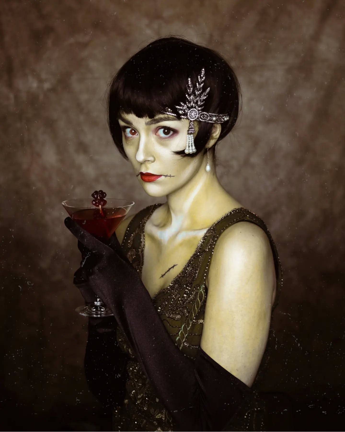 the image is a stylized portrait of a woman dressed in a 1920s flapper dress.  she has a bob haircut adorned with an art deco headpiece and is wearing long black gloves. she holds a cocktail glass containing a dark red liquid. her makeup is carefully applied, giving her a pale, almost ghostly appearance, suggesting a possible halloween costume or a gothic aesthetic. the background is dark and textured, adding to the overall vintage and mysterious mood. the overall style is elegant and retro, evoking the glamour of the roaring twenties. the color palette is muted, with a sepia tone enhancing the vintage feeling.