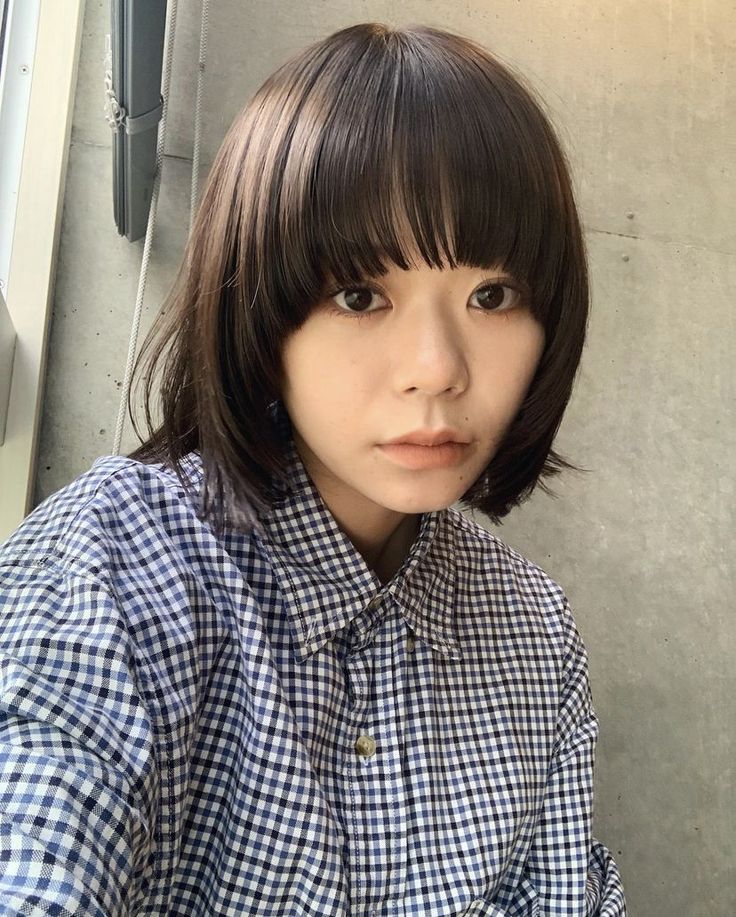a young asian woman with short dark hair and bangs is looking at the camera. she is wearing a blue and white checkered buttonup shirt. the background is a concrete wall.