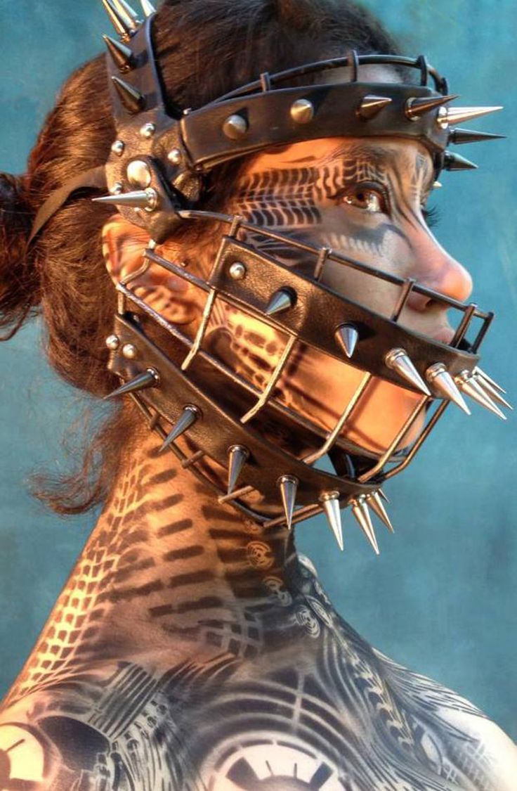 a woman wearing a spiked collar and muzzle. she has a lot of intricate tattoos all over her body, which have a cybergoth vibe. the collar and muzzle are made of leather and metal and have a lot of spikes on them. the image is shot from the front, and the woman is looking to the side. she has dark hair and is wearing a dark top.