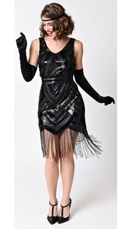 a woman in a black flapper dress with sequins and fringe, wearing long black gloves and a headband. she is posing with her hands on her hips. she has a glamorous look and appears to be ready for a party.