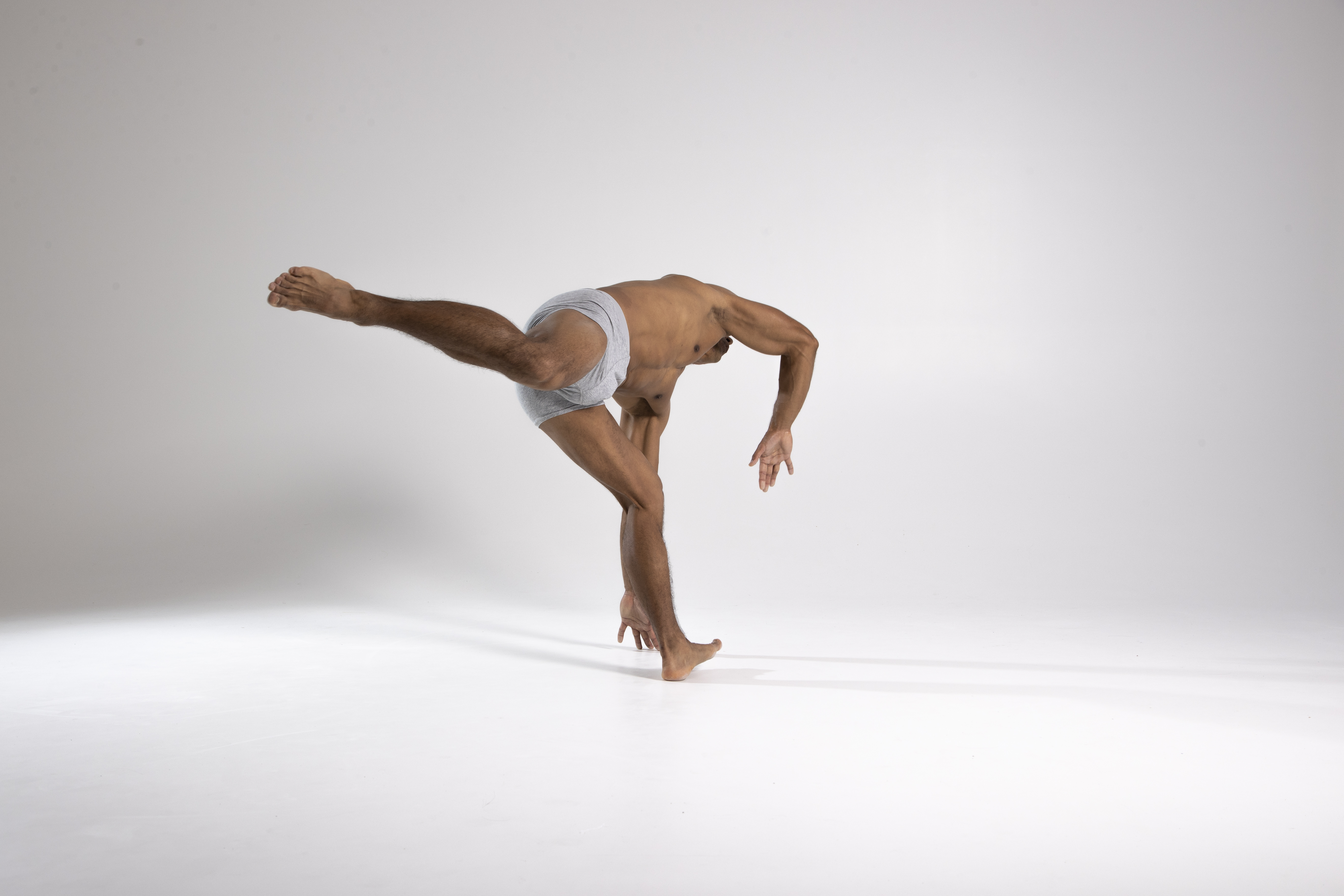 a muscular man stands on one leg, balancing in a modern dance pose, against a white background. the man has toned muscles and wears grey shorts. his arms are extended and his other leg is raised behind him. the lighting is bright, casting a shadow on the floor.