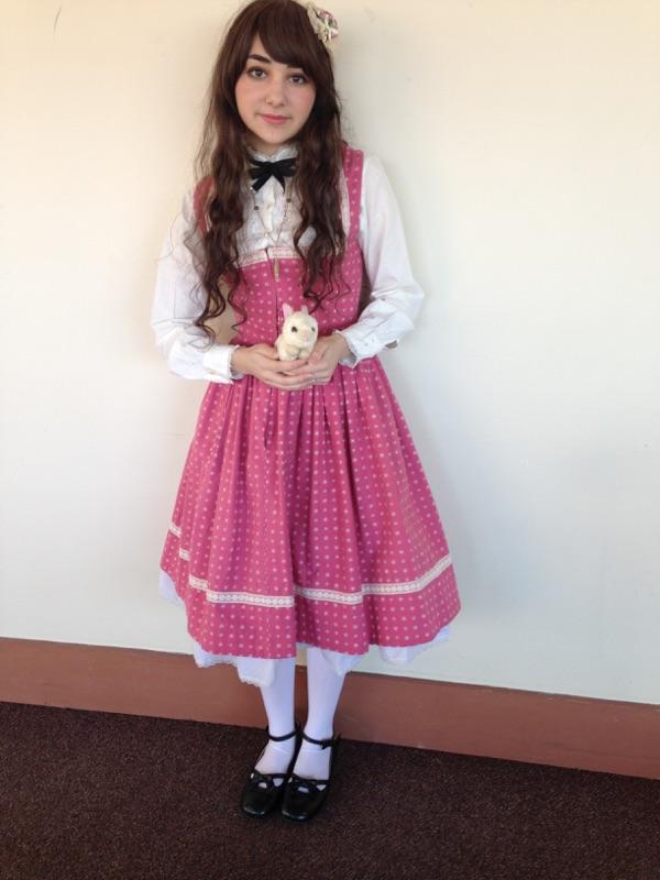 a young girl is standing against a white wall with a brown baseboard. she is wearing a pink polka dot lolitastyle dress over a white blouse. the dress has a full skirt with white lace trim. she is also wearing white socks and black mary jane shoes. she is holding a small white stuffed bunny.  her long brown hair is styled loosely. the overall aesthetic is cute and kawaii.