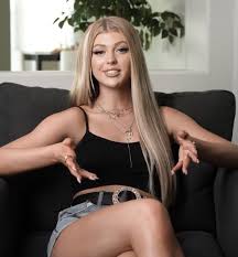 a young woman with blonde hair is sitting on a black couch. she's wearing a black top, jean shorts with a black belt, and layered necklaces. her hands are outstretched. there's a plant behind her, visible through a window or in a frame.