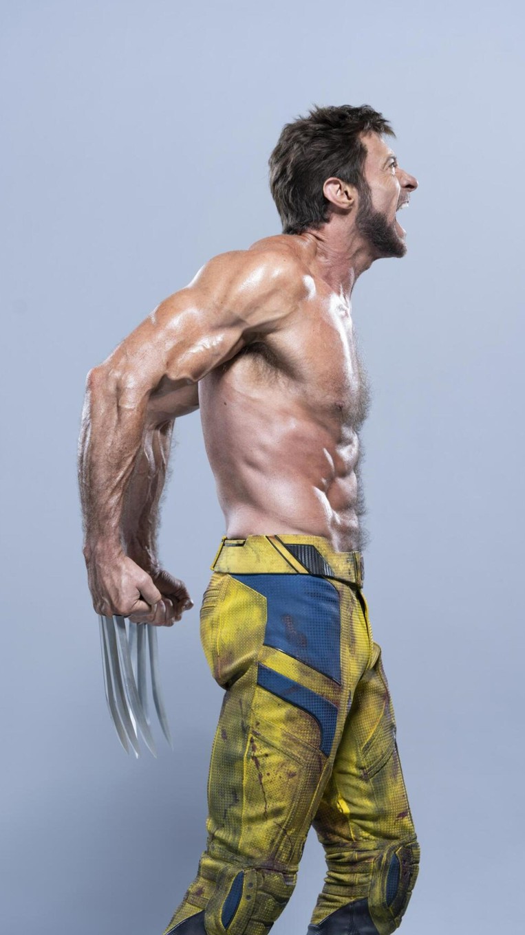 a shirtless hugh jackman as wolverine stands in profile, screaming with his claws extended. he is wearing yellow and blue pants.