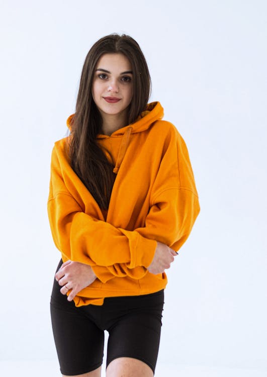 a young woman stands in a studio wearing a bright orange hoodie and black leggings. she is smiling slightly and has long brown hair. she appears to be wearing casual sportswear.