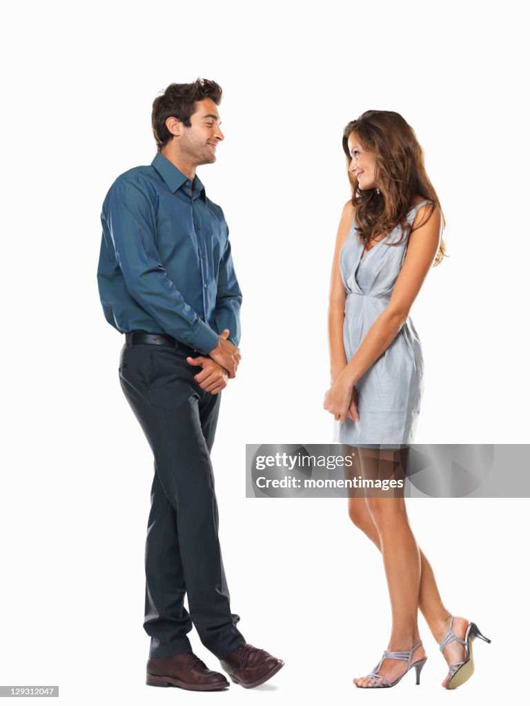 a full studio shot of a man and woman standing opposite each other on a white background, flirting. the man is wearing a dark blue collared shirt, dark gray dress pants and brown dress shoes. the woman is wearing a light blue dress and light gray heels. they are both smiling and looking at each other.