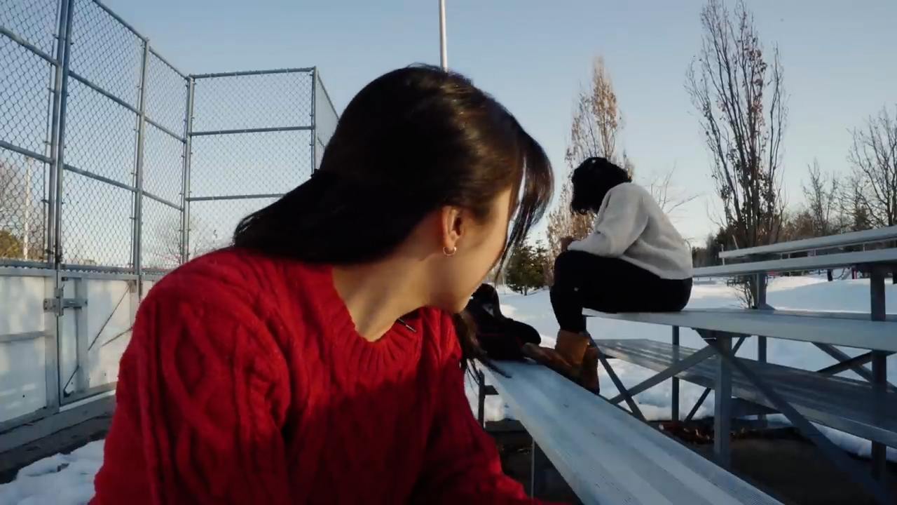three friends are sitting on metal bleachers in the snow on a sunny winter day. the bleachers are located next to a baseball diamond enclosed by a chainlink fence. the perspective is from the point of view of one of the friends sitting on the bleachers.