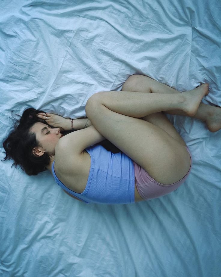 a woman with dark hair is curled up in the fetal position on a white bedsheet. she is wearing a light blue tank top and pink underwear, and she is barefoot. her eyes are closed, and she appears to be sleeping or resting. the overall impression is one of vulnerability, sadness, or introspection.
