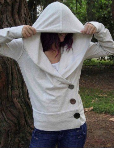 a woman wearing a white hooded sweatshirt with buttons down the front, standing outdoors with a tree trunk in the background. the woman is wearing jeans and has her hood up, covering her head.  she is looking off to the side.