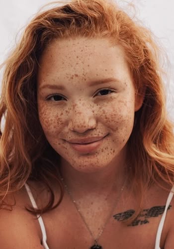 a closeup portrait of a woman with red hair and numerous freckles on her face. she is smiling slightly. she wears a necklace and a white tank top. a bird tattoo is visible on her chest.