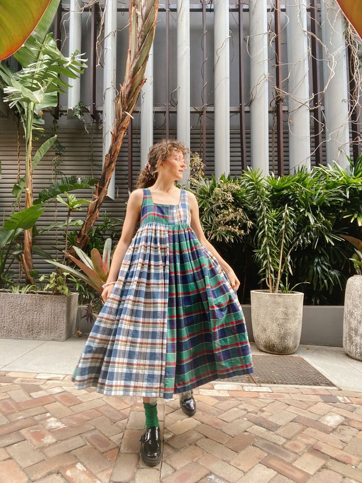 a woman with curly hair stands on a brick patio surrounded by plants, wearing a plaid dress, green socks, and black shoes. she is looking off to the side. the background features plants, a tree trunk, and a building facade with vertical white bars.