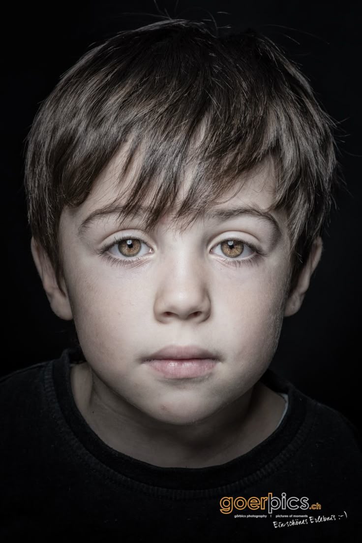 a closeup portrait of a young boy with brown hair and brown eyes against a dark background. the boy has a serious expression. the image includes branding for "goerpics.ch" and "pictures of moments".