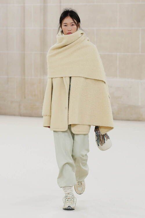 a woman in a white and beige outfit walks down a runway. she is wearing a long, fluffy white coat, a matching scarf, white sneakers, and a small beige bag. she has dark hair and a neutral makeup look. the background is a white wall.