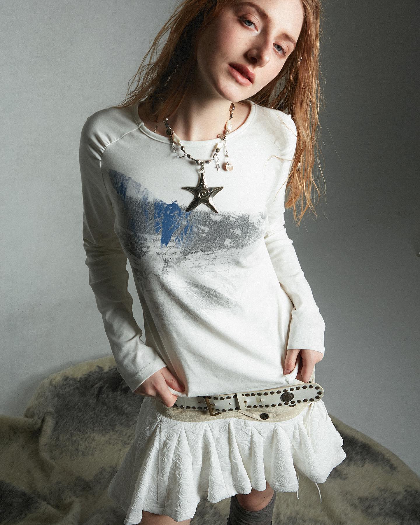 a fullbody shot of a young woman modeling a white longsleeved dress with a landscapeinspired print. she is also wearing a white pleated skirt and a studded belt. her accessories include a necklace with a starfish pendant. her long reddishbrown hair flows loosely around her shoulders.  she's posing against a neutraltoned gray background. the overall style is casual but with a slightly grunge or bohemian feel.
