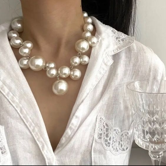 a closeup image of a woman wearing a stylish white pearl necklace. the necklace features large, lustrous pearls arranged in a cascading design. the woman is wearing a white linen shirt with intricate lace detailing. the background is blurred, drawing attention to the necklace and the woman's delicate features. the image conveys a sense of elegance, sophistication, and femininity.