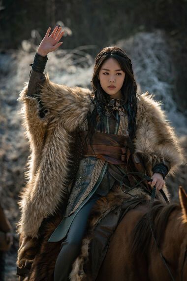a woman with dark hair and fair skin is riding a brown horse. she is wearing a fur cloak over leather armor and has one hand raised in a waving gesture. the background appears to be a natural landscape.