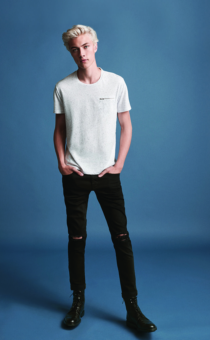 a full body studio shot of a young man with blonde hair, wearing a white tshirt, ripped dark denim jeans, and black boots. he is standing with his hands in his pockets against a blue background.