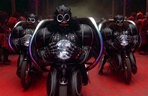 a still from the cult 1979 classic film the warriors featuring three bikers dressed in all black leathers with futuristic helmets and goggles. they are riding their motorcycles and are in the middle of a road, with a red background.