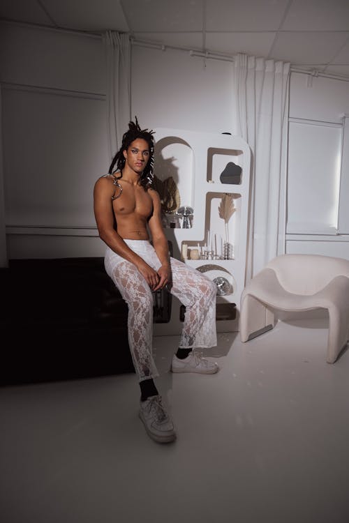 a shirtless male model with dreadlocks sits on a black surface wearing white lace pants and white sneakers. he is posing in a minimalist white studio setting with a white chair and a white shelf in the background. the overall aesthetic is modern and stylish.