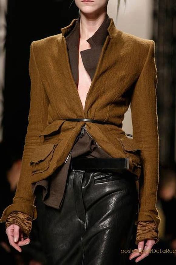 a fashion model in a brown jacket and black leather pants poses on a runway. she has a serious expression and is looking off to the side.