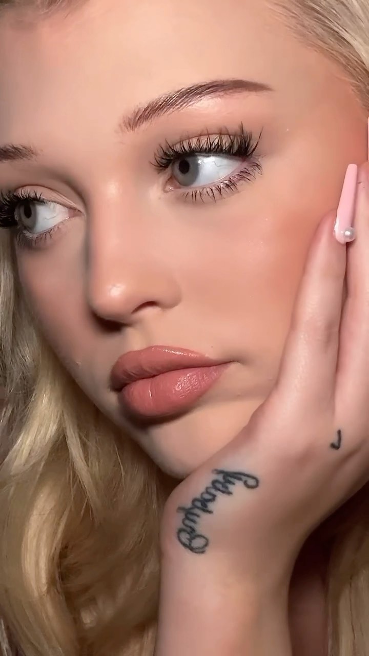 a closeup of a woman's face with blonde hair, makeup, and a tattoo on her hand. her hand is positioned to the right of her face, and she has long eyelashes. her lips are nude colored and shiny.