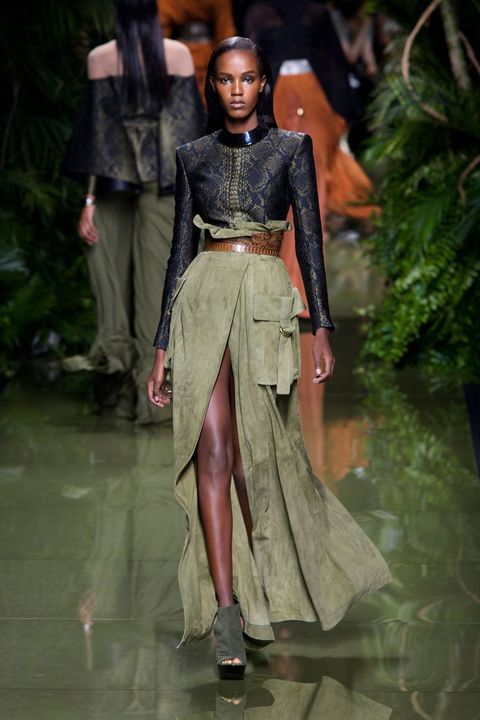 the image shows a darkskinned female model walking down a fashion show runway. she is wearing a stylish outfit consisting of a dark green, snakeskinpatterned cropped top, a long olive green suede skirt with a high waist, a brown leather belt, and dark ankle boots.  the skirt features a thighhigh slit and a cargo pocket detail. the background is blurred but shows lush greenery and other models in similar color palettes suggesting a fashion show setting. the overall aesthetic is sophisticated and highfashion, potentially referencing military or safari themes.  the lighting focuses on the central model.