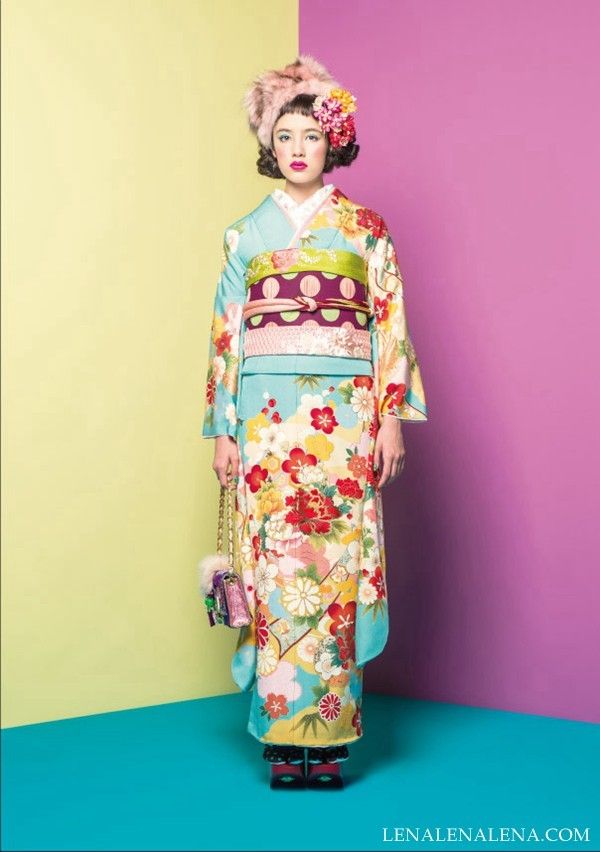 a woman stands in front of a yellow and purple background wearing a light blue kimono with a floral pattern and a fur hat with flowers. she is also carrying a purse. the kimono is tied with an obi. the image is a full shot.