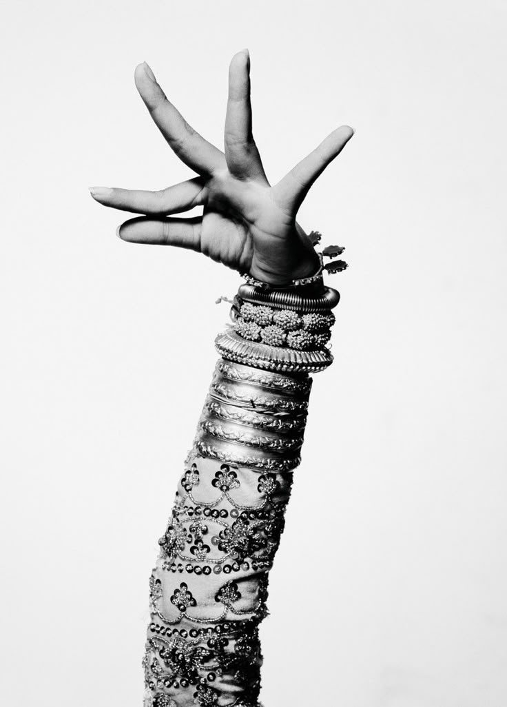 a black and white closeup image of an arm and hand adorned with numerous bracelets, positioned in a mudra gesture, likely associated with indian dance. the sleeve is ornately patterned.