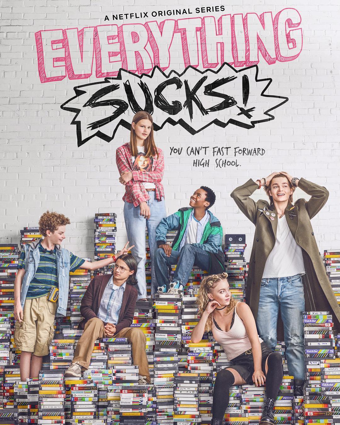the image is a promotional poster for the netflix original series "everything sucks!".  it features a group of teenagers, both male and female, posed around a large stack of vhs tapes. the setting appears to be a brick wall backdrop, lending a retro aesthetic. the title "everything sucks!" is prominently displayed in stylized lettering, along with the tagline "you can't fast forward high school." the overall tone of the poster is nostalgic and evokes the feeling of the 1990s. the characters' clothing and the abundance of vhs tapes solidify the time period and the theme of the show.