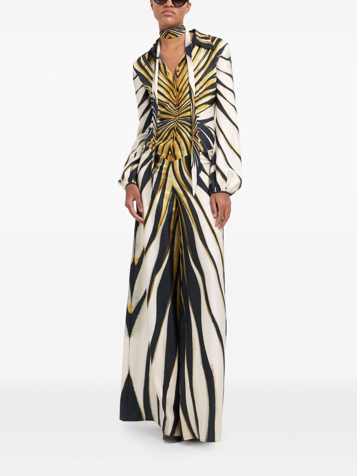a woman is wearing a long, white, black and gold zebra print dress. the dress has long sleeves and a high neckline with a gold scarf tied around it. she is wearing sunglasses and has long hair. the model is standing in a studio with a white background.