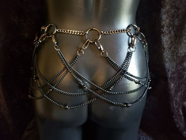the image shows a body chain or waist chain worn on a mannequin. the chain belt is comprised of silver and black metal chains, featuring multiple layers that drape across the lower back and hips. some of the chains also have small beads. the background is a dark, textured surface.