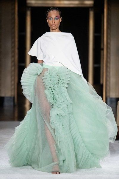a fashion model walks down a runway in a  green tulle skirt with ruffled details.  the skirt is paired with a white, simple top.  the model has dark hair and smokey eye makeup.