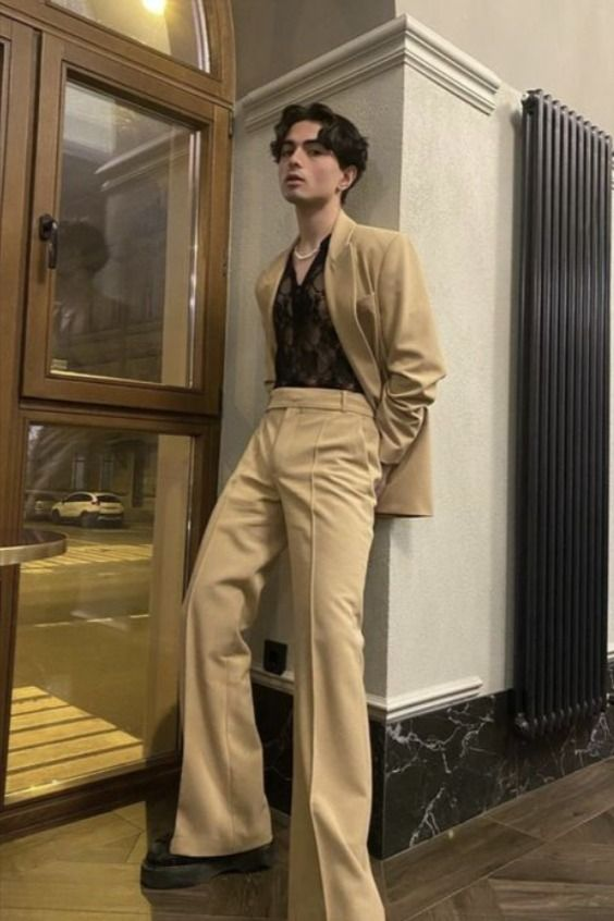 a young man stands in front of a window in a trendy brown suit jacket and pants with a black lace shirt, looking dapper and stylish.  he is wearing black shoes and  leaning on a creamcolored wall. the background shows wood floors and a brown wall.