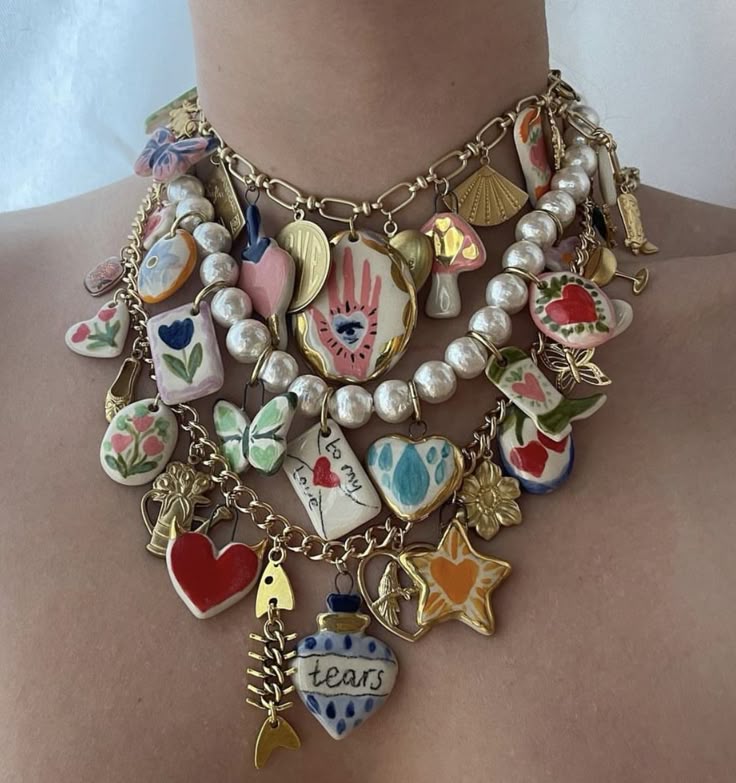 a closeup shot of a person wearing a layered charm necklace. the necklace consists of a gold chain and a pearl necklace, both laden with various colorful and whimsical charms. the charms are a mix of ceramic pendants with handpainted designs and goldcolored metal charms. some of the prominent charm designs include hearts, stars, butterflies, flowers, and other unique shapes. the overall style of the necklace is playful and kitsch. the person's skin is visible in the background.