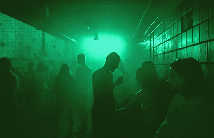 the image depicts a dimly lit nightclub or warehouse party scene. the predominant color is a dark, hazy green, created by green lighting.  numerous people are present, but their features are indistinguishable due to the low light and smoke or haze. only silhouettes are visible, creating a sense of anonymity and a mysterious atmosphere. the setting appears industrial, possibly a converted warehouse space, with metal grids visible on the walls. the overall mood is one of energy and clandestine activity.