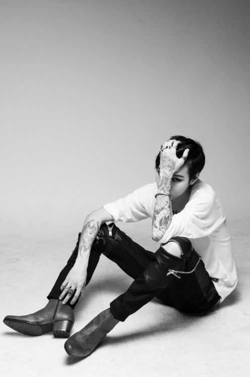 a young man with tattoos on both arms sits on the floor with his legs crossed. he is wearing a white shirt, ripped jeans, and boots. he is holding his hand to his head and looking down at the ground. the photo is taken in a studio and is in black and white.