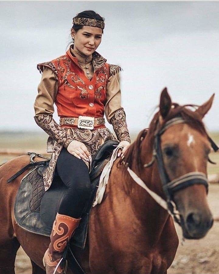 a woman in a traditional outfit is sitting on a brown horse, wearing a headband, and looking directly at the camera. the horse is facing the camera and the woman is riding in a natural setting with a wideopen field. the image is taken in daylight and the woman appears to be enjoying horseback riding.
