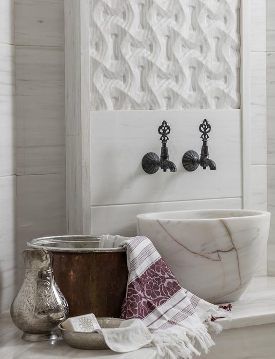this is an image of a luxurious turkish bath, also known as a hammam. it features a white marble wall with an intricately carved geometric pattern. two ornate, dark bronze faucets are mounted on the wall, above a large, white marble sink. a fluffy white towel with a purple floral pattern is draped over the edge of the sink. a copper bucket with ornate silver details sits on the counter to the left, next to a bar of soap on a silver tray. the scene exudes tranquility and relaxation.