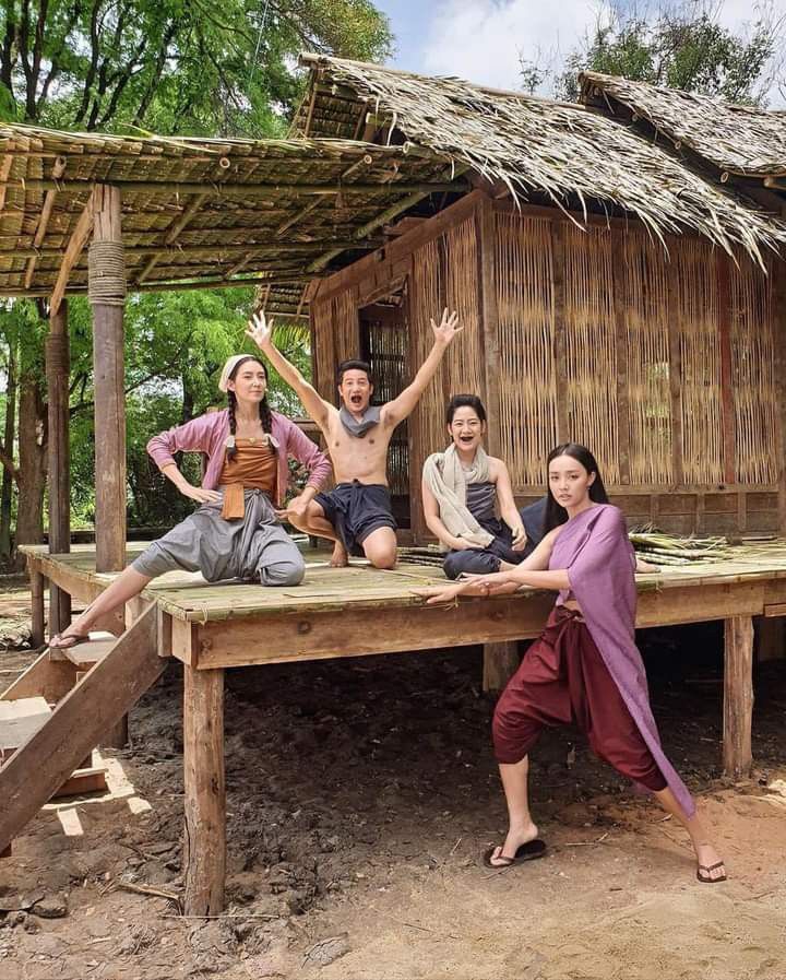 this photo shows four people standing and sitting in front of a traditional wooden house in thailand. they are all smiling and looking happy. the three women are wearing traditional thai clothing while the man is shirtless. they appear to be having a good time and enjoying their time together.  the photo was taken outdoors, and the setting is a rural one, with a dirt path in the foreground. the background shows a lush green forest, giving a sense of peace and tranquility.