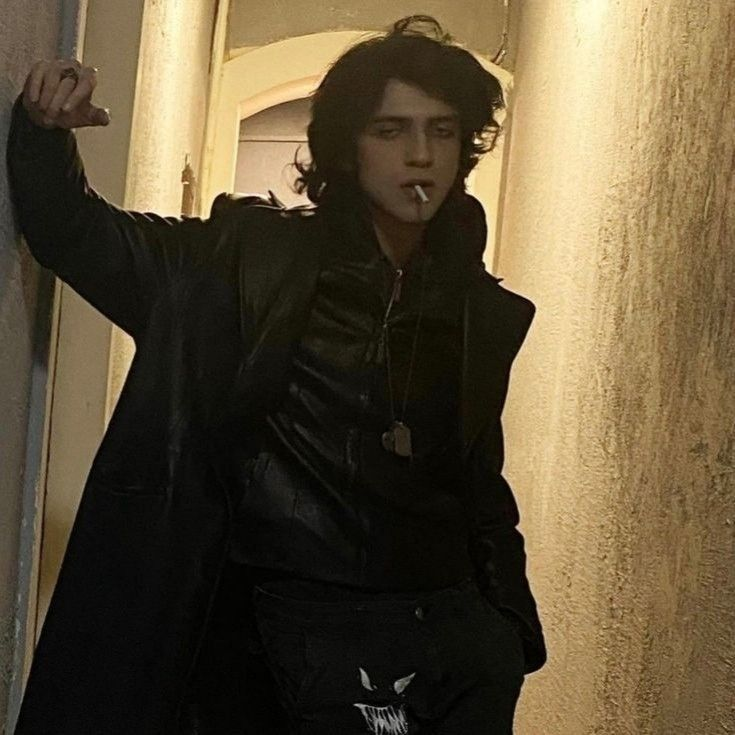 a young man wearing a black leather jacket and black jeans is standing in a doorway. he has long black hair and is smoking a cigarette.