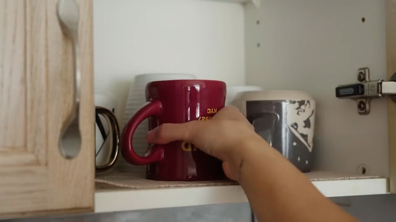 a woman's hand reaches for a red mug inside a kitchen cabinet. the cabinet door is open, revealing a shelf lined with a tan mat and holding an assortment of mugs.  the cabinet has a light wooden finish, and the wall it's mounted on is white.
