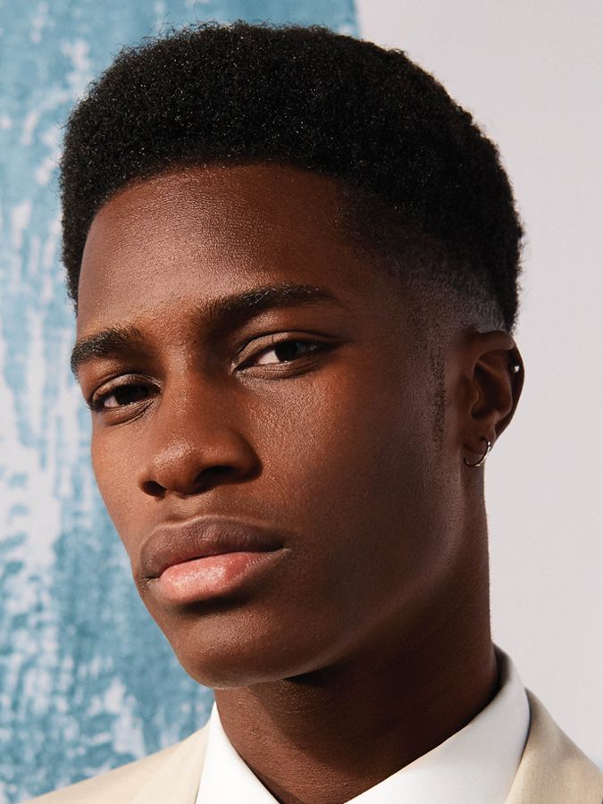 closeup portrait of a young black man with short, textured afro hair styled with a fade. he is wearing a beige suit and white shirt and has small hoop earrings in his ears.  his expression is serious and intense. the background is a blurred, light teal and white pattern. the image focuses on his face and upper body.
