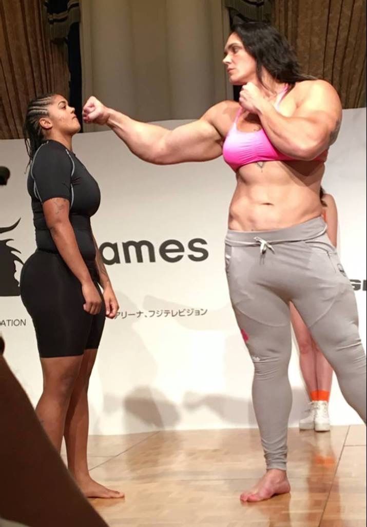 a picture of two female athletes standing side by side. the taller female athlete is much larger and more muscular than the smaller one. the larger woman is wearing grey pants and a pink sports bra. the smaller woman is wearing black pants and a black shirt. they are both posing for a photo.