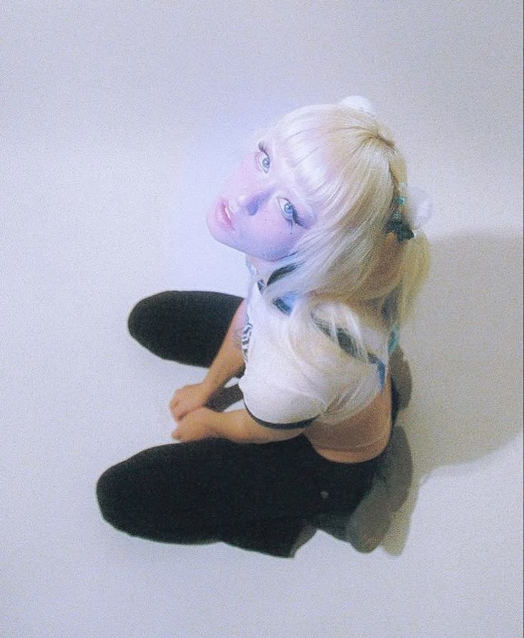 a woman with platinum blonde hair and blue eyes kneels and looks up. she is wearing a white tshirt, black pants, and head accessories. she has fair skin and blue makeup.