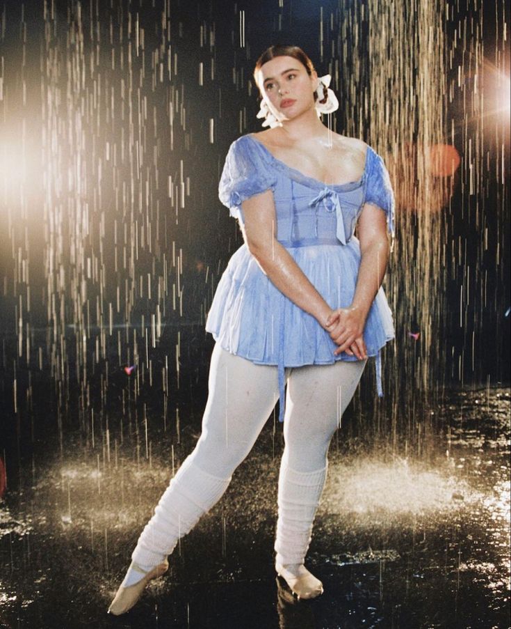 the image shows a plussize woman, styled as a ballerina, standing in the rain. she's wearing a light blue, shortsleeved, balletstyle dress with a ruffled hemline, white tights, and white balletstyle footwear. the background features a continuous sheet of artificial rain falling. the overall mood is romantic and artistic, with the lighting playing off the water droplets and highlighting her dress. the model's pose is graceful and elegant, emphasizing her figure in a bodypositive manner. the image has a professional photoshoot aesthetic.