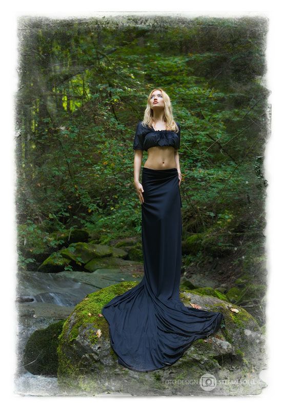 a blonde woman in a black crop top and long, flowing skirt stands on a mosscovered rock in the middle of a forest stream. she is looking up towards the sky, and her skirt spreads out around her on the rock. the forest is lush and green, and the stream is flowing over rocks in the foreground.